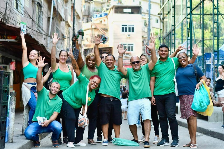 Marcelinho das Comunidades com a equipe da campanha, todos de verde e com os braços erguidos, numa rua de comunidade no Rio.